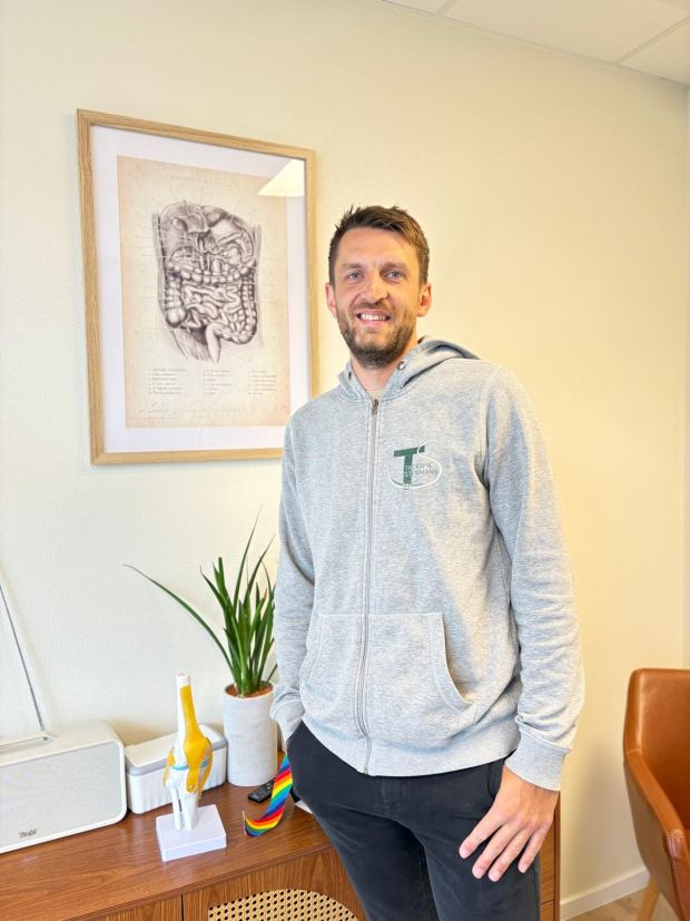 Thomas Stermann in seiner Praxis, in grauer Sweatjacke mit dem Logo der Therapie Stermann; dahinter eine gerahmte anatomische Zeichnung und ein Modell eines Kniegelenks.